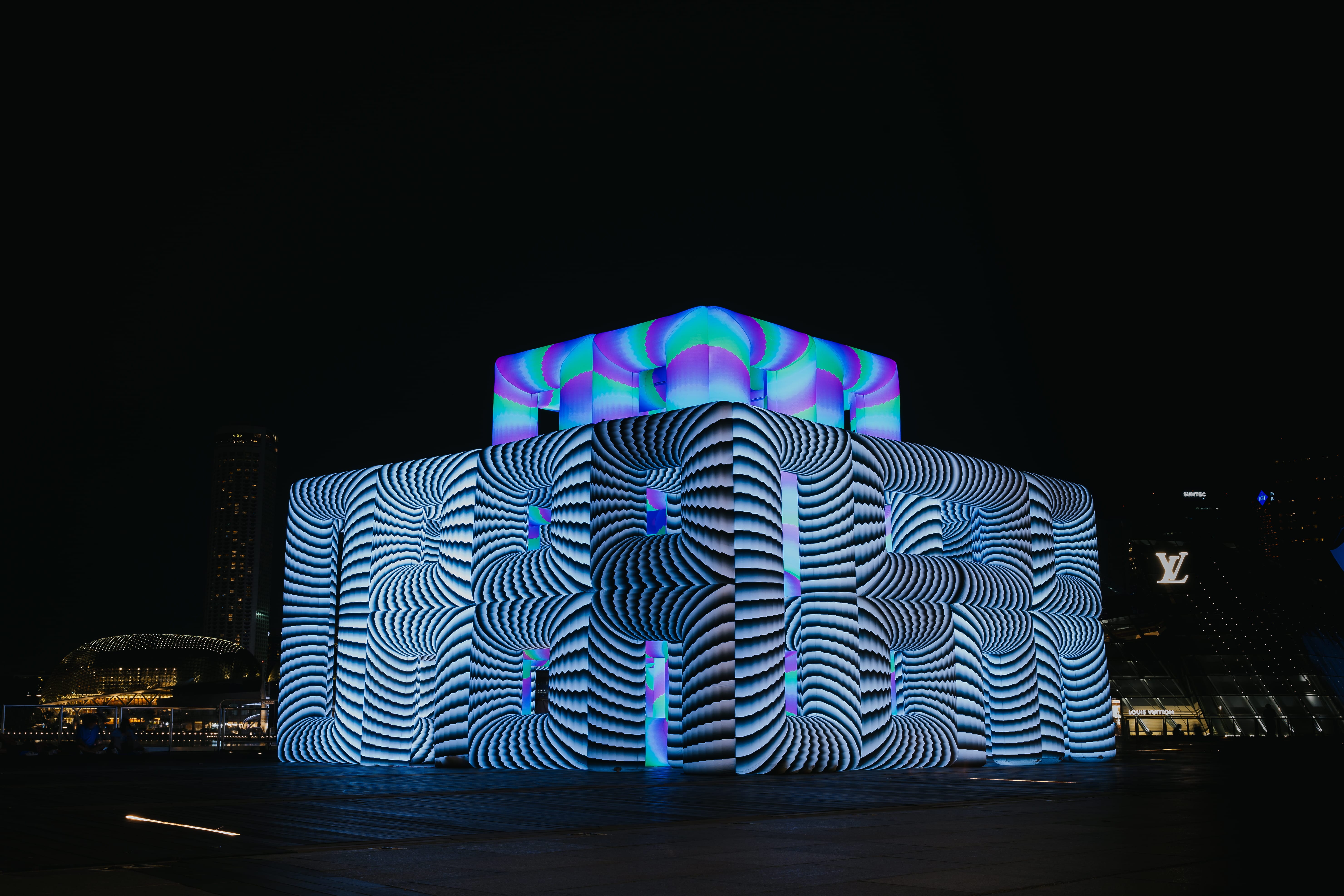 A large cube-shaped light art installation covered in swirling black-and-white patterns, topped with vibrant pink and green projections, with visitors standing around it at night.